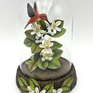 Puede incluir: Una cúpula de cristal que contiene un colibrí esculpido en naranja y negro sobre flores de cítricos blancas con hojas verdes. La base es de resina texturizada con aspecto de madera marrón, decorada con flores blancas y hojas verdes.
