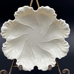 May include: A cream-coloured ceramic dish with a scalloped, ruffled edge and a central embossed floral pattern featuring six radiating stems that extend toward the rim, displayed on a decorative bronze-toned metal wire stand.