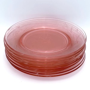May include: A stack of six pink glass plates. Each plate has a delicate etched leaf design around the rim. The plates are a soft, translucent pink color and appear to be vintage or antique.