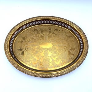 May include: An oval gold-toned metal tray featuring an intricate engraved floral pattern on the base. The tray has a raised, openwork lattice border and a textured, rope-style rim, providing a polished finish to the metallic surface.