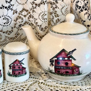 May include: A cream-colored ceramic teapot and two salt and pepper shakers. Each piece features a red birdhouse design with a bird perched on top. The teapot has a handle and spout. The set is trimmed with a blue-green border.