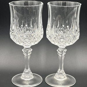 May include: Two clear crystal wine glasses with diamond-cut detailing on the bowl and stem. The glasses have a classic shape with a wide base and a flared rim. The stems are intricately designed, and the glasses are set against a dark background.