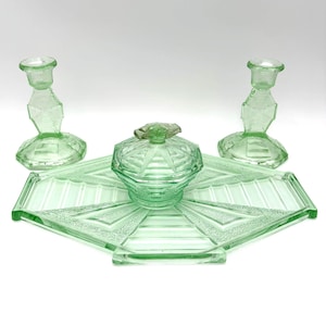 May include: A set of light green pressed glass items including two matching candlesticks, a lidded bowl, and a large hexagonal tray. Each piece features a geometric pattern with textured and smooth surfaces, creating a cohesive glass collection.