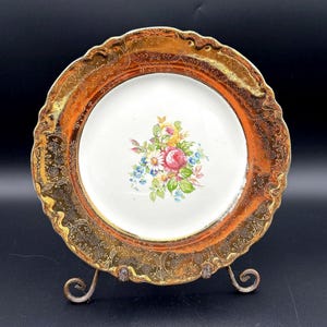 May include: A vintage decorative plate with a gold rim and floral center. The plate features a white center with a bouquet of pink, yellow, and blue flowers. The gold rim has an ornate, embossed design. The plate is displayed on a decorative metal stand.