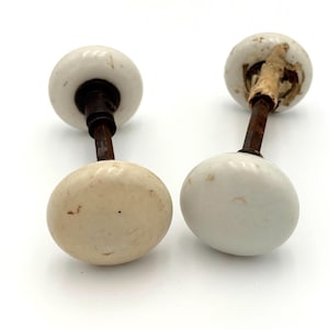 May include: Two antique white ceramic door knobs with aged metal stems. The knobs have a rounded shape and show signs of wear, suggesting they are vintage. The door knobs are set against a white background.