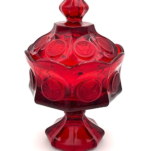 Puede incluir: Un bombonera rojo rubí con tapa. El recipiente de vidrio tiene una forma geométrica y una base de pedestal. La tapa está adornada con diseños circulares en relieve. Una pieza vintage que añade elegancia.