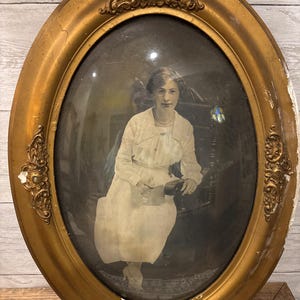 Puede incluir: Un marco de fotos antiguo de forma ovalada con un acabado dorado. El marco tiene detalles ornamentados y una cubierta de vidrio. Una fotografía en blanco y negro de una mujer con un vestido blanco está dentro del marco.