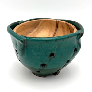 May include: A handmade, two-piece bowl. The outer bowl is a teal-green ceramic with several small holes and a rough texture. The inner bowl is made of light-colored wood with a natural grain pattern. The bowl has a rustic, organic aesthetic.