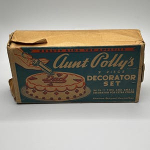 Op de afbeelding: Een kartonnen doos met een blauw en wit etiket waarop "Aunt Colly's 9 Piece Decorator Set" staat. Het etiket bevat ook een afbeelding van een taart die is versierd met rode glazuur en het woord "Dad".