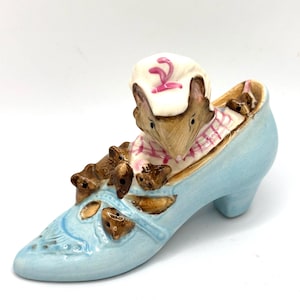 May include: A light blue ceramic shoe figurine with a mouse wearing a chef's hat and a pink and white plaid shirt. The shoe has brown accents and cut-out details.