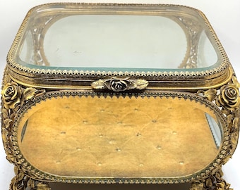May include: An antique gold-toned jewelry box with a clear glass top and an oval mirror base. The box features ornate floral detailing, including roses on the corners and a rose clasp. The interior base has a quilted pattern.