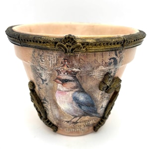 Puede incluir: Maceta decorativa con diseño de pájaro. La maceta está pintada en color melocotón claro con un acabado envejecido. Un pájaro con corona aparece en la parte delantera. El borde y los detalles decorativos son dorados.