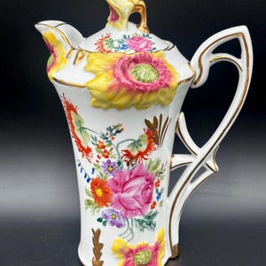 May include: A white porcelain teapot featuring raised, three-dimensional yellow and pink floral relief patterns. The body is decorated with painted floral bouquets in shades of pink, orange, red, and purple, accented with gold trim on the handle, spout, and lid.