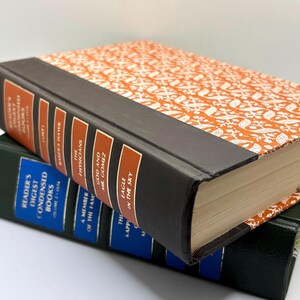 May include: Two stacked Reader's Digest Condensed Books. The top book has a dark brown spine with orange labels and an orange cover with a white floral pattern. The bottom book has a dark green spine with blue labels and white text.