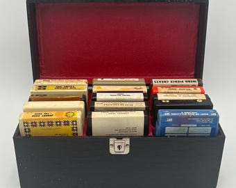May include: A black, rectangular cassette tape storage box with a red interior. The box is filled with various cassette tapes, some labeled with artist names like Johnny Cash and Webb Pierce. The box has a silver clasp and handle.