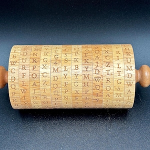 Puede incluir: Una rueda de cifrado de madera con letras y números grabados. El cuerpo cilíndrico tiene anillos giratorios, cada uno con un conjunto diferente de caracteres. Las perillas de madera están en cada extremo para girar. La rueda es de madera clara.