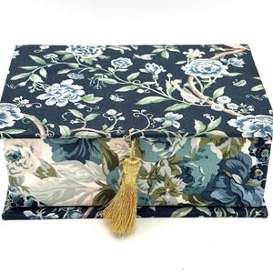 May include: A rectangular jewelry box with a floral pattern in shades of blue, green, and white on a dark blue background. The box has a gold tassel attached to the front. The floral design includes flowers and leaves.