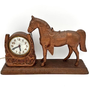 May include: A bronze-colored horse clock with a horseshoe-shaped clock face. The clock features a horse statue with a saddle. The clock face has black numbers and hands. The base has a decorative border.