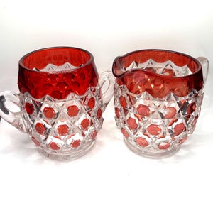 May include: A clear glass creamer and sugar bowl set with a red rim and diamond-cut pattern. The creamer has a handle and a spout, while the sugar bowl has a handle. Both pieces are on a clear base.
