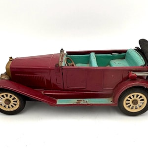 May include: A vintage burgundy toy car with a convertible top. The car features cream-colored wheels, a teal interior, and a black convertible top. The car has a detailed front grill and headlights.