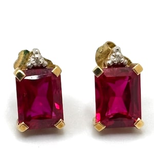 May include: A pair of stud earrings featuring rectangular, deep pink faceted stones set in gold-toned metal. Each earring is accented with a small, triangular silver-toned detail at the top, which is set with three tiny, clear, sparkling stones.