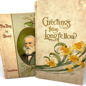 Op de afbeelding: Twee antieke boeken met versleten covers. Eén boek leest "The Day is Done" en de andere "Greetings from Longfellow". Het tweede boek bevat gele bloemenillustraties. Beide boeken hebben een portret van een man.