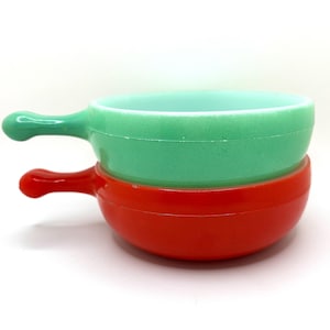 Peut inclure: Deux bols ronds empilés avec poignées, l'un vert clair et l'autre rouge vif. Chaque bol présente une finition lisse de couleur unie et une seule poignée saillante sur le côté. Les bols sont placés l'un sur l'autre sur un fond blanc.