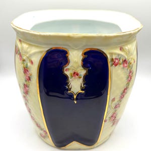 May include: A vintage ceramic planter with a cream-colored background, adorned with pink floral patterns and gold trim. The planter features two large, dark blue panels with gold outlines, adding a striking contrast to the design. The top rim is a light blue color.