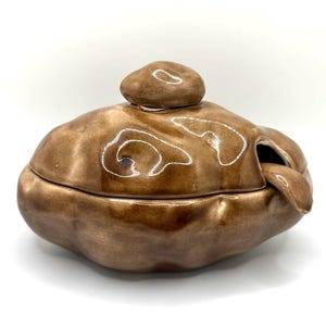 May include: A brown ceramic lidded bowl with an irregular, organic shape and a glossy finish. The lid features a rounded knob handle, and a matching ceramic spoon rests inside the bowl, protruding through a small opening on the side of the rim.