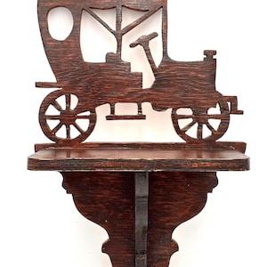 May include: A dark brown wooden shelf in the shape of an antique car. The shelf features a detailed cutout design of the car's body, wheels, and driver's area. The shelf is mounted on a decorative bracket.