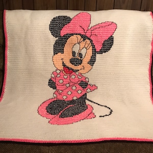 May include: A white crocheted blanket with a pink and black Minnie Mouse design. The blanket has a pink border.