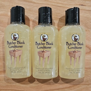 May include: Three bottles of Howard Butcher Block Conditioner, each with a black cap and a clear body. The labels feature a logo and text, including "Food Grade Mineral Oil & Natural Waxes" and "2 FL. OZ. (59 ml.)". The bottles are arranged on a wooden surface.