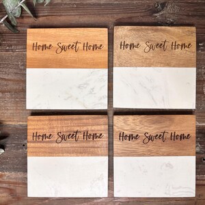 May include: A set of four square coasters featuring a two-tone design with natural brown wood on the top half and white marble on the bottom half. Each coaster is engraved with the black cursive text Home Sweet Home across the wooden section.