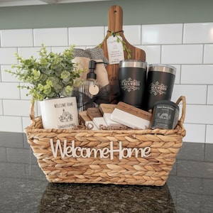 May include: A woven basket with the text Welcome Home containing a faux plant in a white pot, a glass soap dispenser, a wooden cutting board, two black tumblers labeled Blackwood, a small candle, and a set of white and wood coasters.