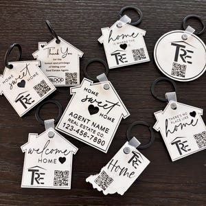 May include: A collection of silver-colored keychains in various shapes, including houses and a circle. Each keychain features the text "Home Sweet Home," "Welcome Home," or "Home is Where the Heart Is." Some include a QR code and the "TRE" logo.
