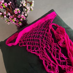May include: A bright pink, open-weave net bag with a matching handle. The bag is made of a woven material and is displayed on a dark green surface. A vase of pink and white flowers is in the background.