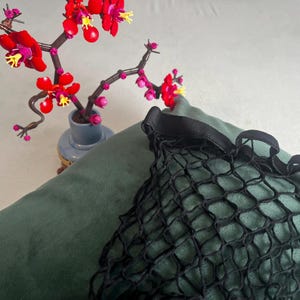 May include: A still life featuring a dark green pillow and a black net bag. A small blue vase holds a decorative branch with red flowers and yellow accents. The scene is set against a neutral background.