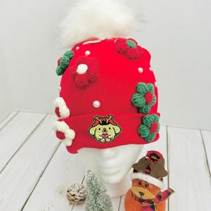 May include: A red knit beanie with white faux fur pom-pom, adorned with green and red crocheted flowers and white pearls. The beanie features an embroidered cartoon character. A festive winter hat.