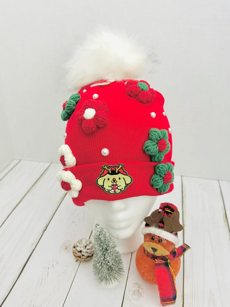 May include: A red knit beanie with white faux fur pom-pom, adorned with green and red crocheted flowers and white pearls. The beanie features an embroidered cartoon character. A festive winter hat.