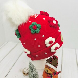 May include: A red knit hat adorned with white faux fur pom-pom, green and red floral embellishments, and small white pearls. The hat is decorated with white and red flowers. A small Christmas tree and a festive ornament are in the foreground.