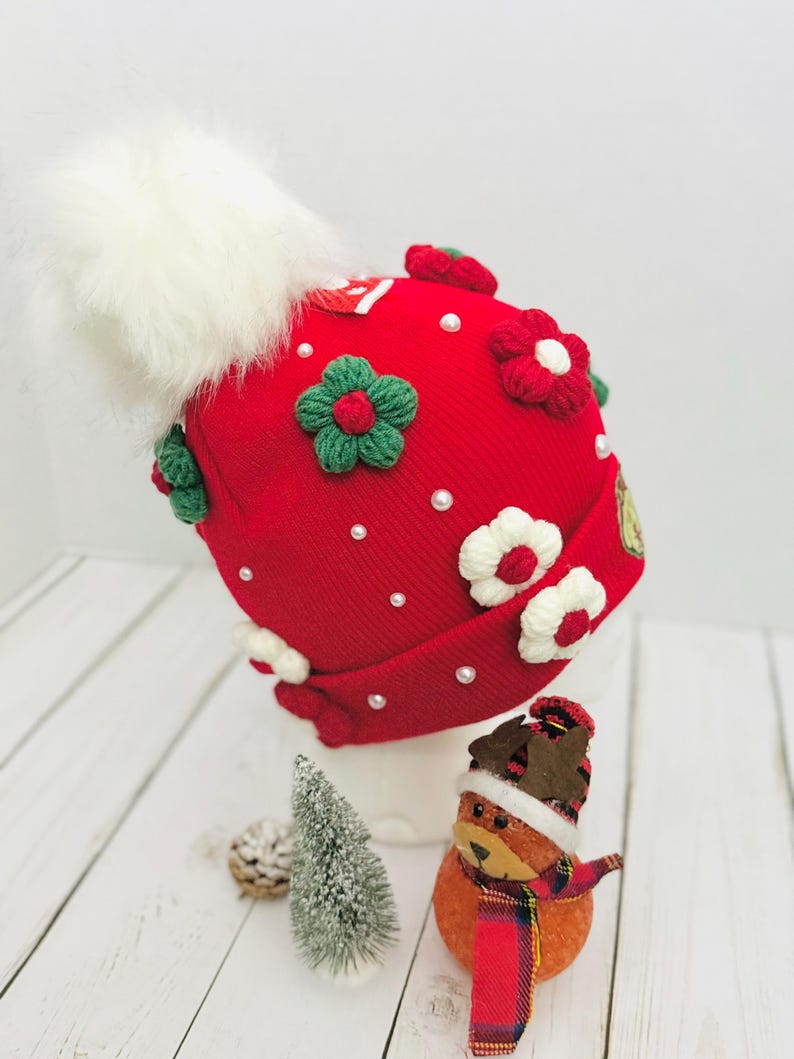May include: A red knit hat adorned with white faux fur pom-pom, green and red floral embellishments, and small white pearls. The hat is decorated with white and red flowers. A small Christmas tree and a festive ornament are in the foreground.