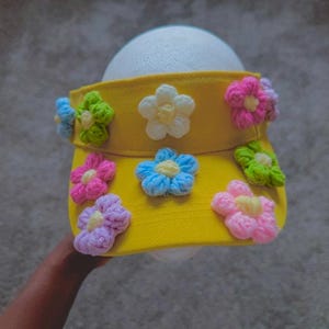 Puede incluir: Visera amarilla adornada con flores de ganchillo coloridas. Las flores son rosas, azules, verdes y blancas, a&ntilde;adiendo un toque l&uacute;dico. La visera est&aacute; dise&ntilde;ada para proteger los ojos del sol.