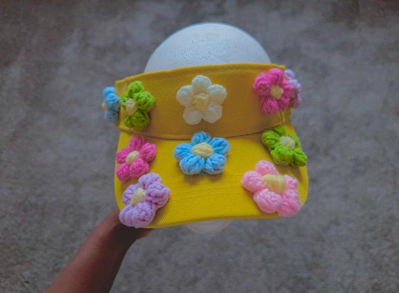 Puede incluir: Visera amarilla adornada con flores de ganchillo coloridas. Las flores son rosas, azules, verdes y blancas, a&ntilde;adiendo un toque l&uacute;dico. La visera est&aacute; dise&ntilde;ada para proteger los ojos del sol.
