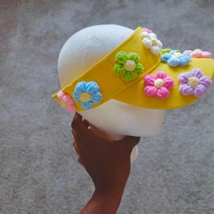 Puede incluir: Una visera amarilla adornada con flores bordadas de colores, rosas, azules, verdes y moradas. La visera est&aacute; sujeta por una cabeza de espuma blanca, mostrando el dise&ntilde;o floral.