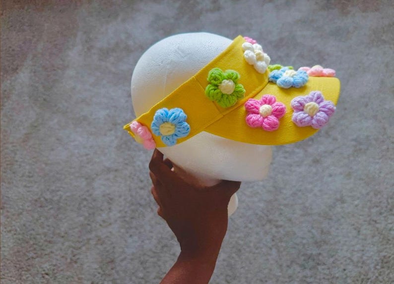 Puede incluir: Una visera amarilla adornada con flores bordadas de colores, rosas, azules, verdes y moradas. La visera est&aacute; sujeta por una cabeza de espuma blanca, mostrando el dise&ntilde;o floral.
