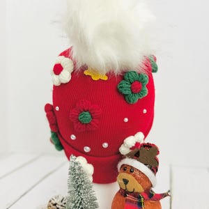 May include: A red knit hat with white faux fur and floral embellishments. The hat is decorated with white, red, and green crocheted flowers and small pearl accents. A small Christmas tree and a bear figurine with a scarf are in the foreground.