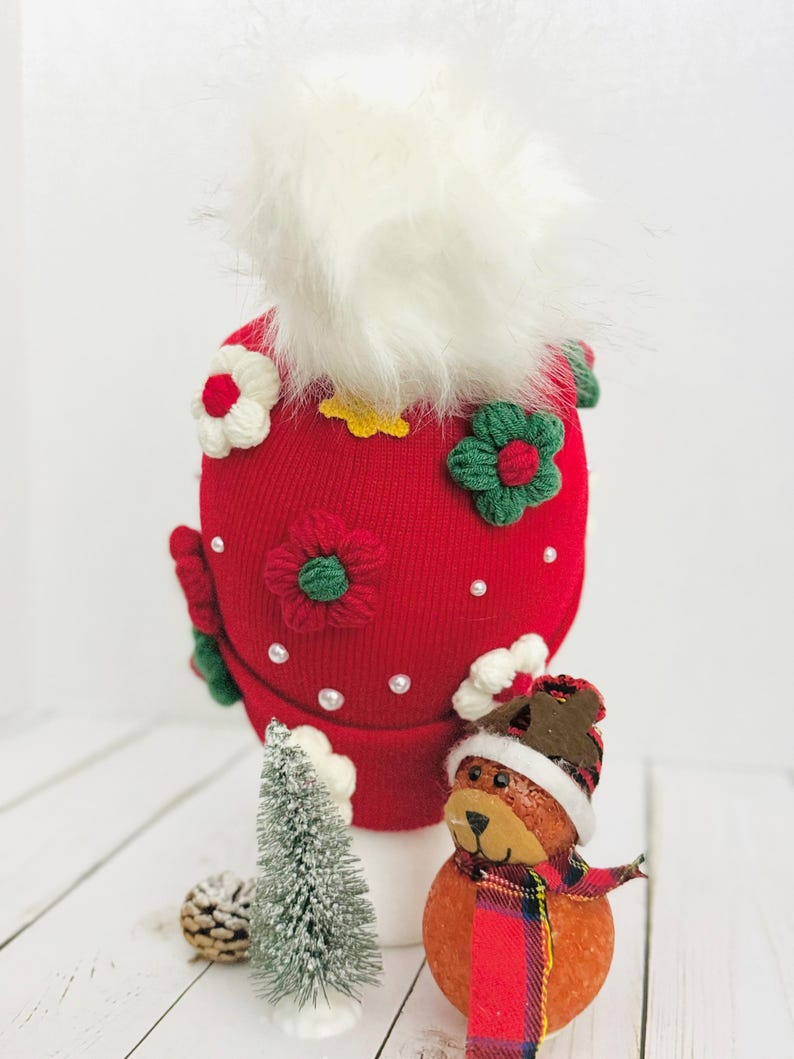 May include: A red knit hat with white faux fur and floral embellishments. The hat is decorated with white, red, and green crocheted flowers and small pearl accents. A small Christmas tree and a bear figurine with a scarf are in the foreground.