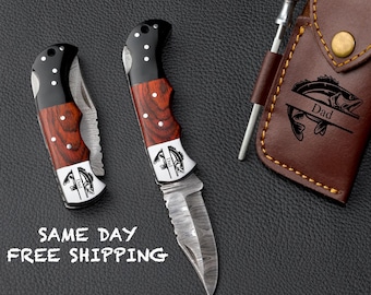 May include: Two pocket knives with black and red handles and a fish design engraved on the blade. The knives are on a dark gray surface. The knife on the right has the word "Dad" engraved on the handle. The knives come with a brown leather sheath.