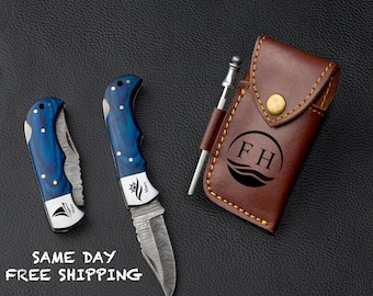 May include: Two folding knives with blue wood handles and patterned blades, alongside a brown leather case with a gold snap and the initials "FH". A silver pen is also included. The knives have a nautical emblem.