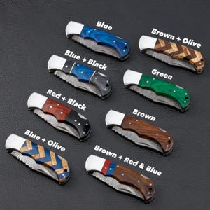 Collection of handcrafted Damascus steel folding knives with various custom wood handle designs including 
blue, green, chevron, and mixed wood patterns. Compact and durable pocket knives ideal for personalized graduation gifts, 
outdoor use, or groomsmen sets.